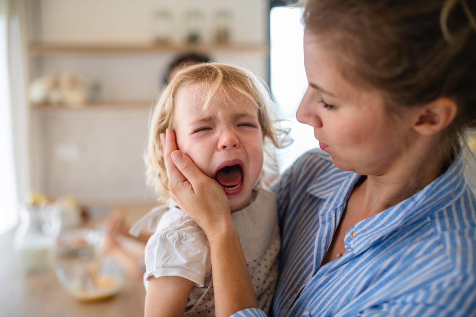 A Mother Holding A Crying Toddler Daughter Indoors In Kitchen.